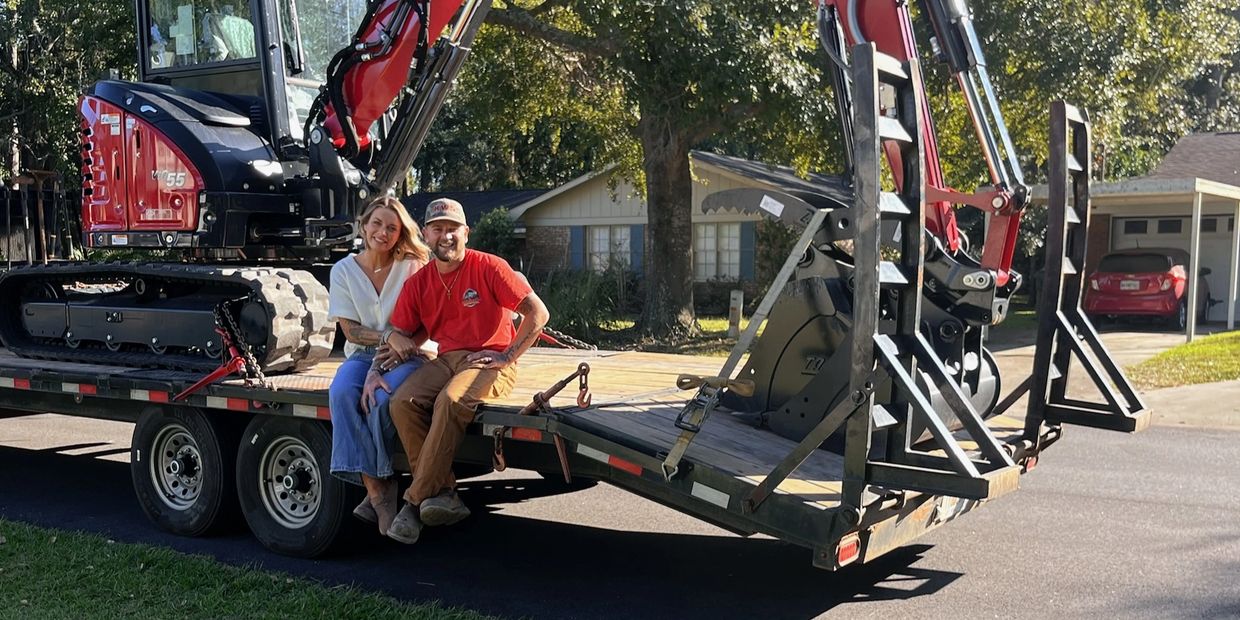 A couple sitting on a flatbed trailer with a red excavator behind them.