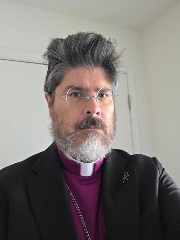 Portrait of a man wearing clerical attire with glasses and a gray beard.