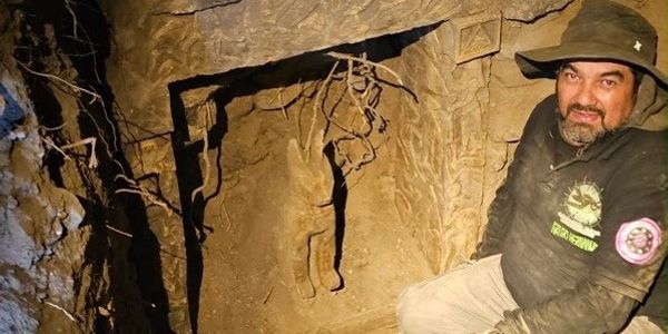 Man in a hat next to a carved wall showing a hanging figure in a cave.