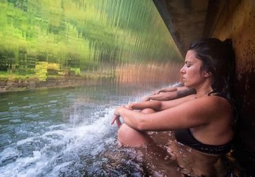 Woman in black bikini sitting relaxed under a waterfall indoors.