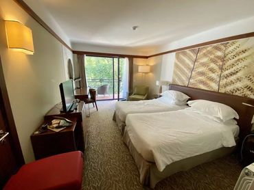 Cozy hotel room with twin beds and balcony overlooking greenery.