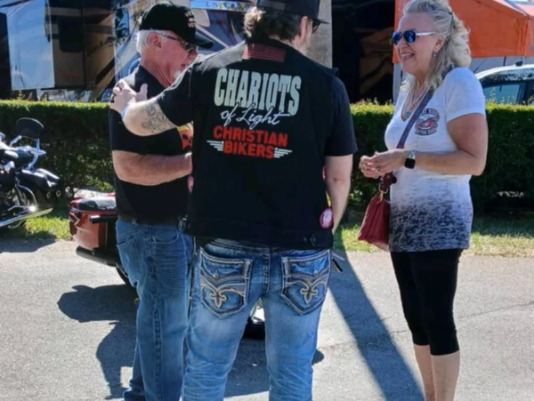 Three people conversing outdoors on a sunny day with motorcycles nearby.