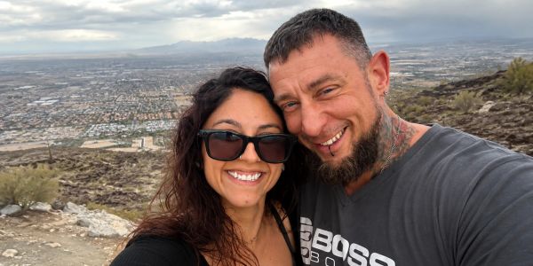 A happy couple takes a selfie on a mountain with a cityscape background.
