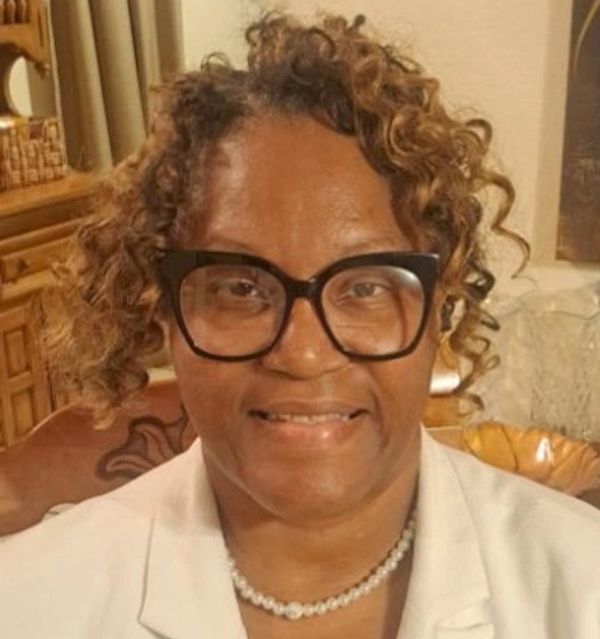 Smiling woman with curly hair, glasses, and pearl necklace.