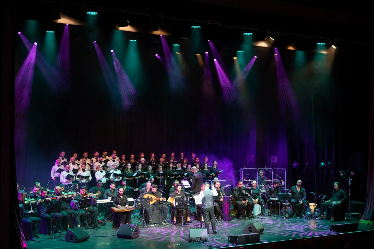 A large orchestra and choir perform on a colorful lit stage.