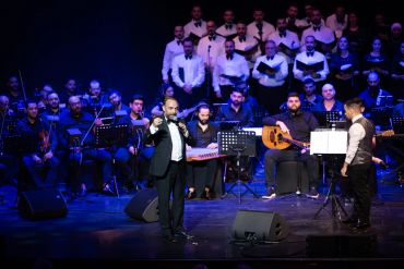 Toni Bayeh speaking on stage with orchestra and choir in the background.