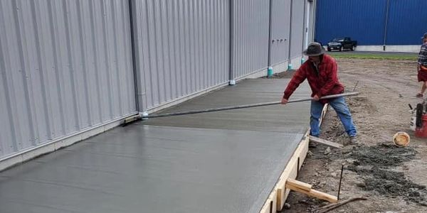 Worker finishing freshly poured commercial concrete slab alongside metal building in Massachusetts.