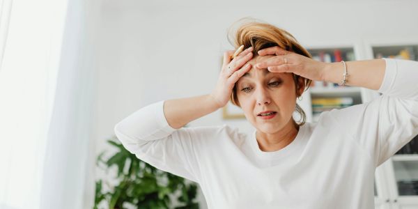 Woman in white shirt looks stressed, holding her forehead indoors.