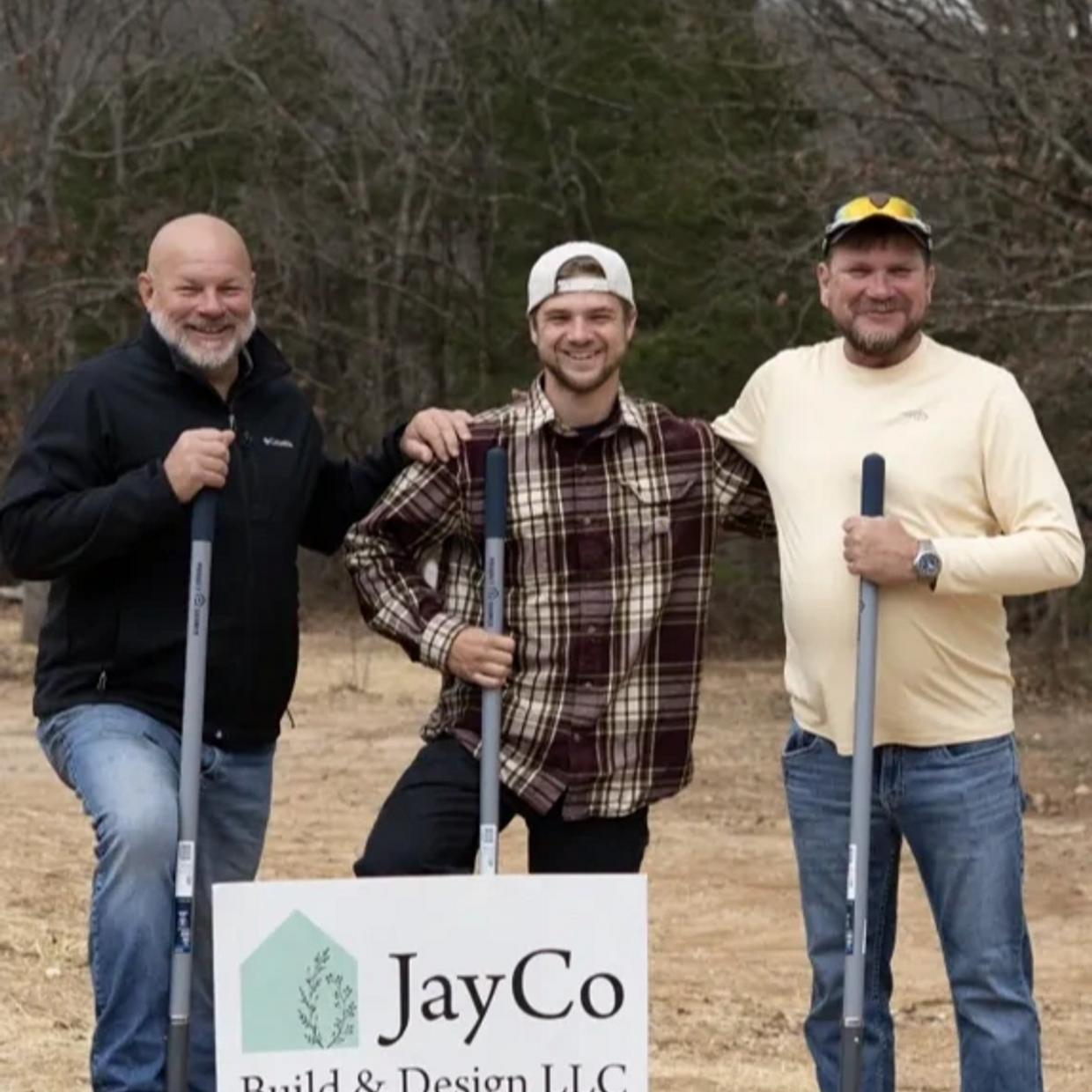 Three men smiling outdoors with a JayCo Build & Design LLC sign and shovels.