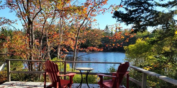 Two red chairs and a small table on a deck overlooking a serene lake surrounded by autumn trees.