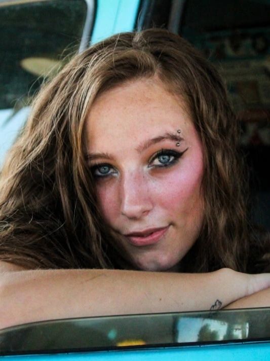 Young woman with blue eyes and eyebrow piercings leans on a car window.