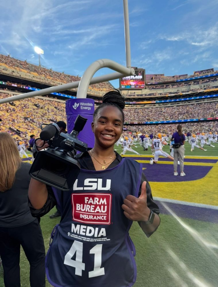 A smiling female camerawoman at an LSU football game giving a thumbs up.