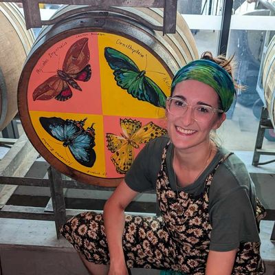 A smiling woman in floral overalls and a headband poses by a colorful butterfly painting.