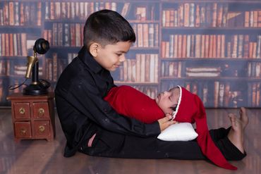 Older boy tenderly holds a sleeping baby wrapped in red with a long hat.