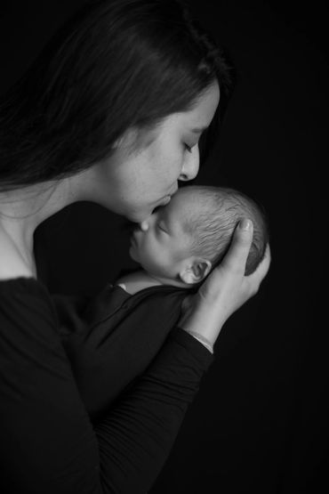 A mother tenderly kisses her newborn baby in a black and white portrait.