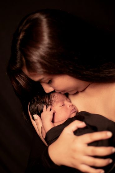 A mother tenderly embraces and kisses her newborn baby wrapped in a black cloth.
