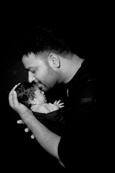 A father tenderly holds and kisses his newborn baby in a black and white portrait.