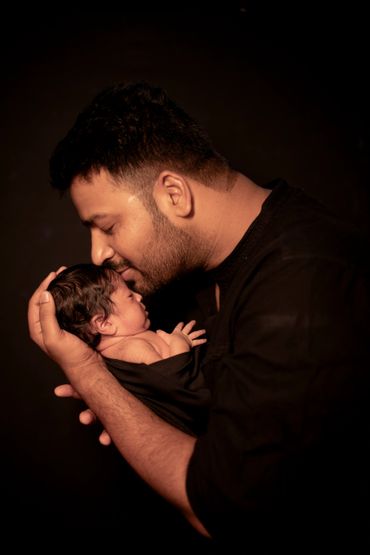 A man gently holds and kisses a newborn baby, wrapped in black cloth.