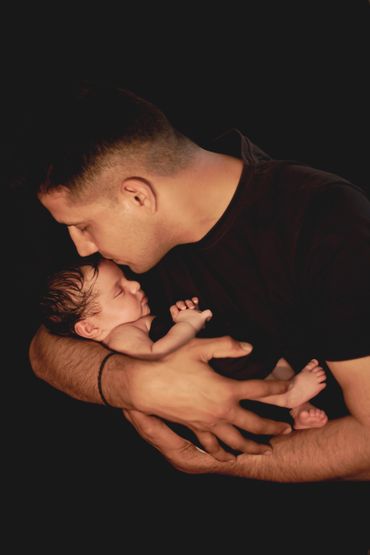 A man tenderly kisses a sleeping baby held in his arms against a dark background.