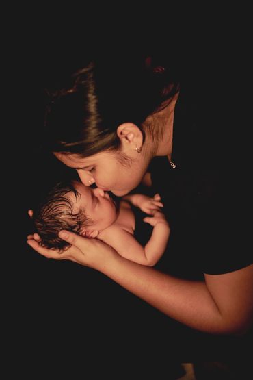 A mother lovingly kisses her newborn baby against a dark background.