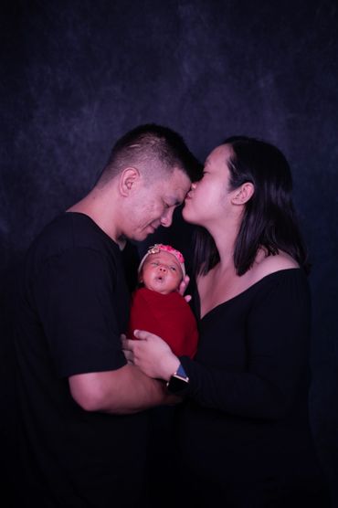 Mother kissing father while holding newborn baby wrapped in red.