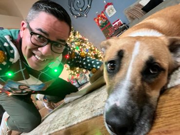 Man in festive sweater smiles next to a dog lying on the floor near a decorated Christmas tree.