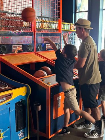 A boy shoots a basketball in an arcade game while a man watches.