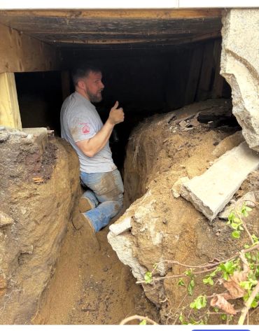 Man giving thumbs up while kneeling in a muddy trench inside a wooden structure.