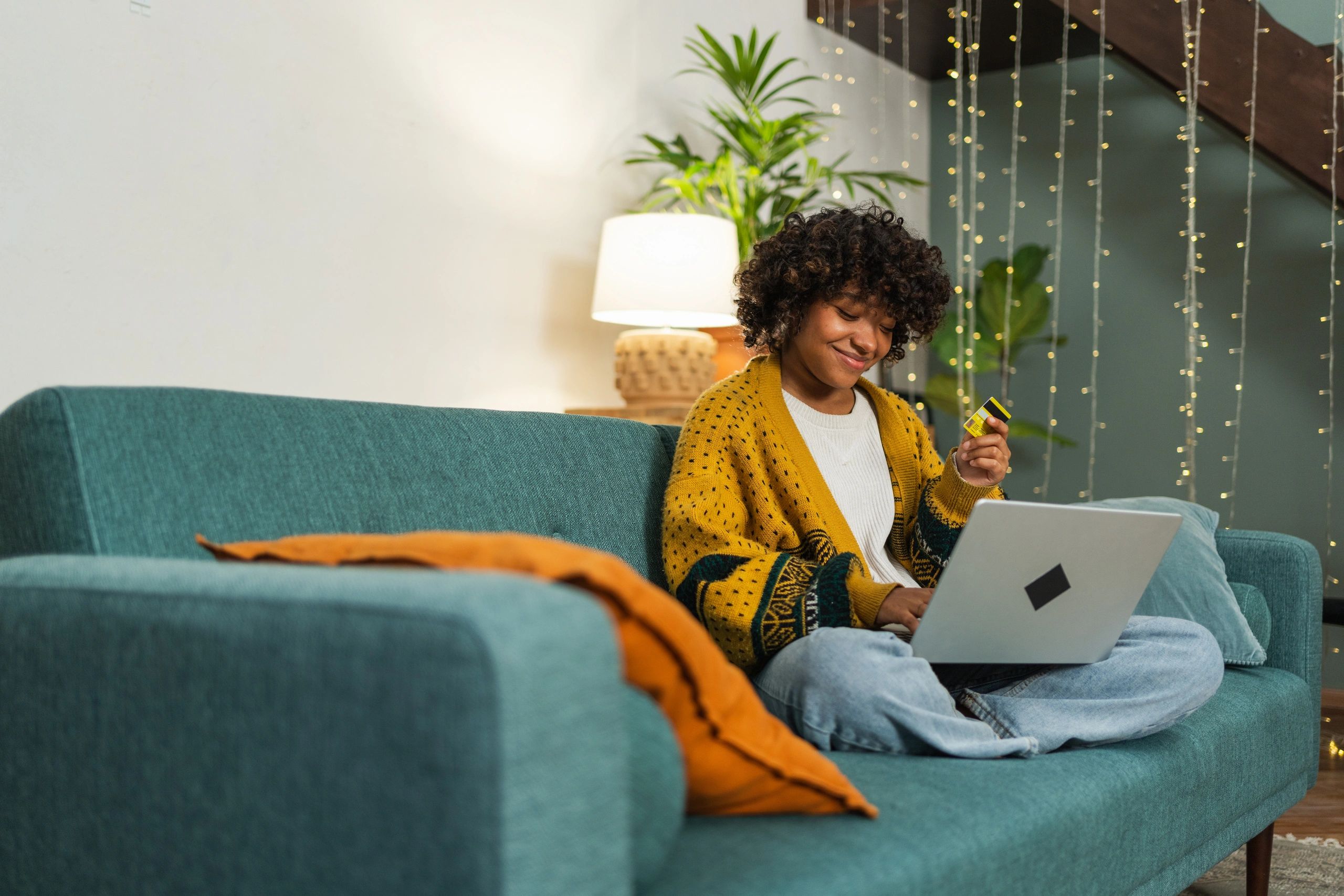 A woman dressed in a yellow cardigan on a sofa with a laptop on her knees and a card in her hand