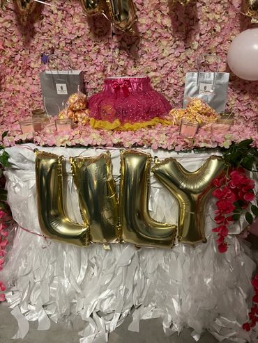 A decorative party table with pink flowers, gold balloons spelling "LILY", and party favors.