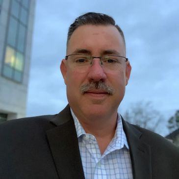 A man in glasses, suit jacket, and checkered shirt outdoors with a building and cloudy sky.
