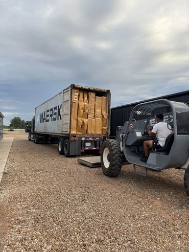 A man operates a forklift near a truck loaded with boxes on a gravel path.