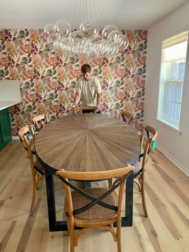 Man adjusting chairs around a round wooden dining table in a floral wallpapered room.