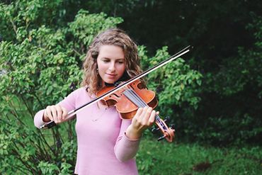 A woman playing violin outdoors surrounded by greenery.