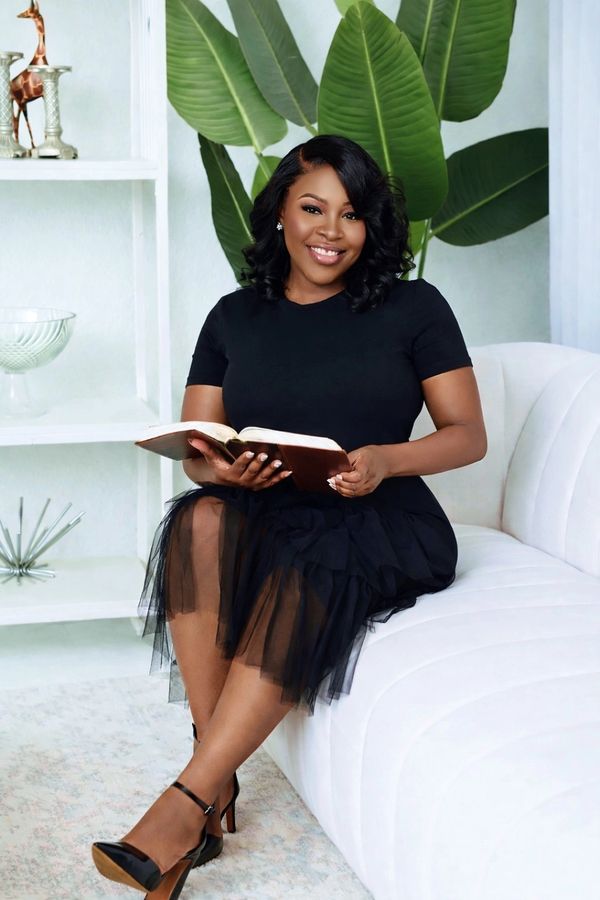 Woman in black dress sitting on white couch, smiling with a book in hand.