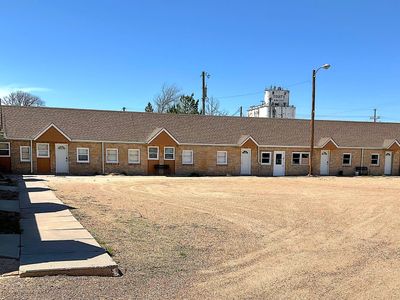 A row of rustic motel rooms under clear blue sky with a large gravel parking area.