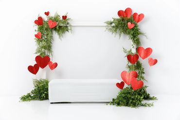 Valentine’s Day mini session set up, large white frame with greenery and red hearts on the corners.
