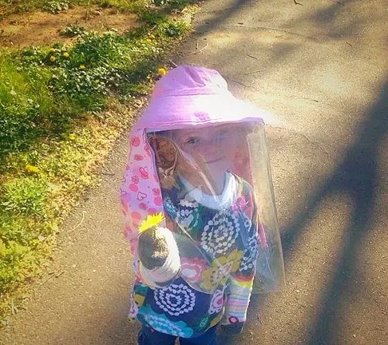 Child wearing a pink hat with a face shield holding a flower.