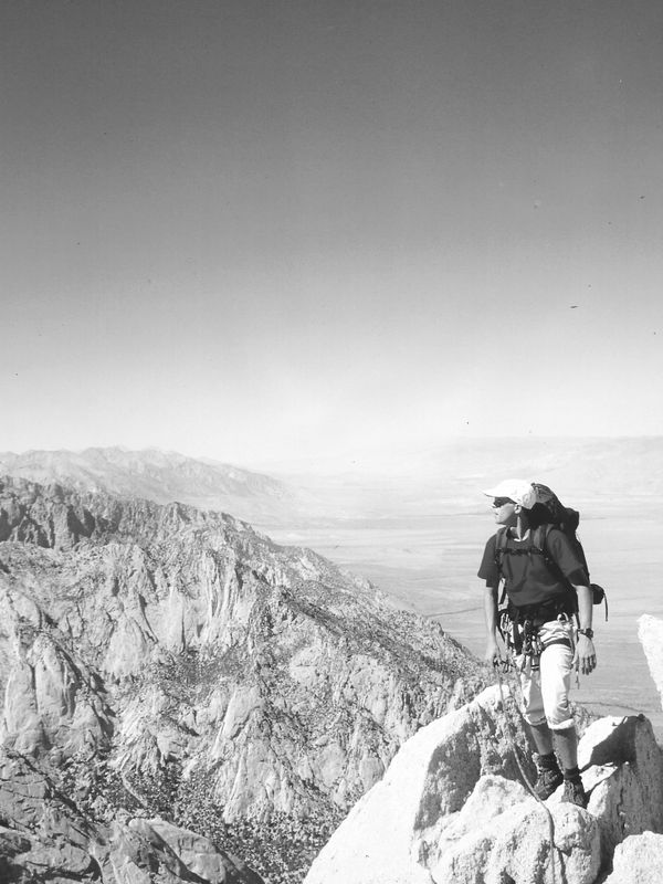 A climber stands on a rocky peak overlooking vast mountain ranges under a clear blue sky.