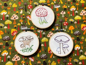 Three embroidery hoops with mushroom designs on colorful mushroom-patterned fabric.