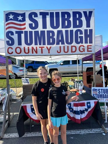 Two girls smiling in front of a Stubby Stumbaugh County Judge campaign booth.