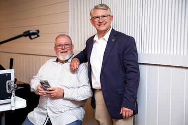 Two older men smiling, one seated with a phone, the other standing in a blazer.