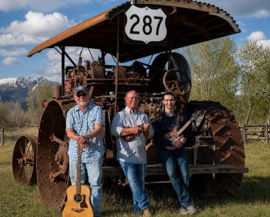 Three men with string instruments posing in front of an old rusty tractor under a highway sign.