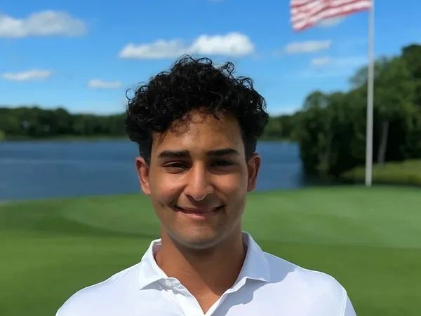 Young man smiling outdoors with a golf course and American flag in the background.