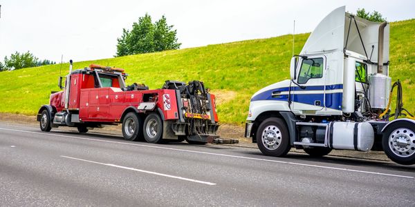 A red tow truck hooking up a white semi-truck on the roadside.