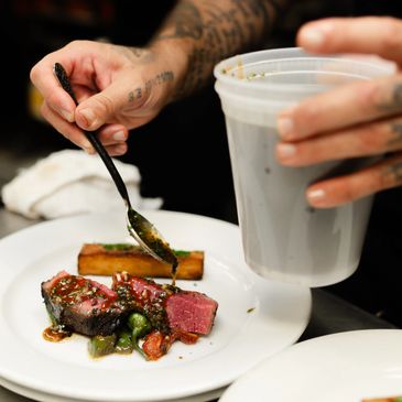 Chef carefully drizzles sauce over seared steak with vegetables on a white plate.