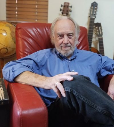 Elderly man sitting comfortably in a red leather chair, with guitars and a globe in the background.