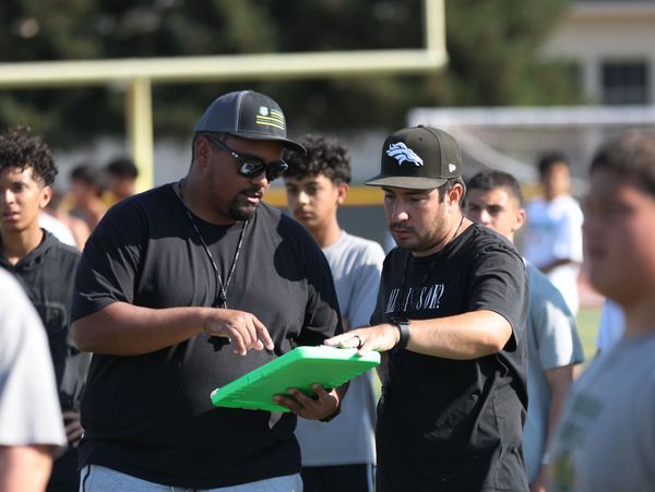 Two coaches discuss strategy on a football field with players around them.