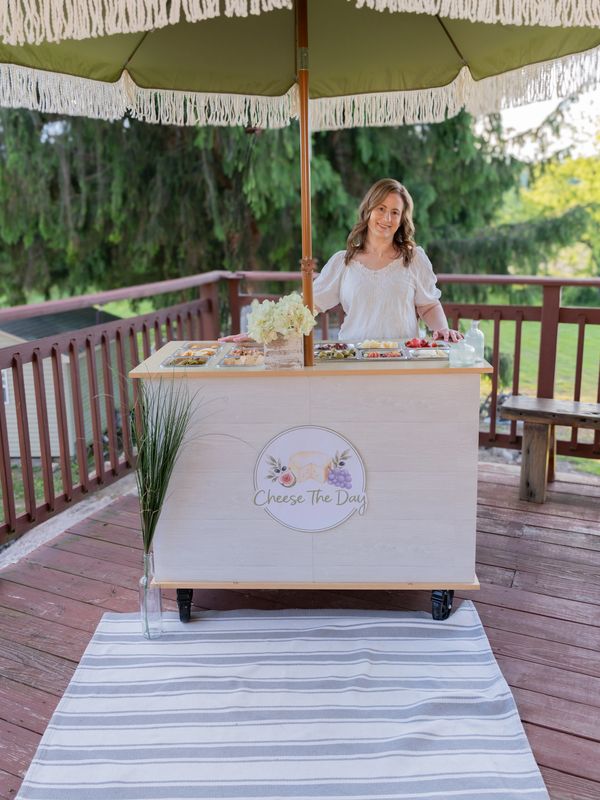 Woman standing behind a charcuterie bar cart under a green umbrella serving cheese.