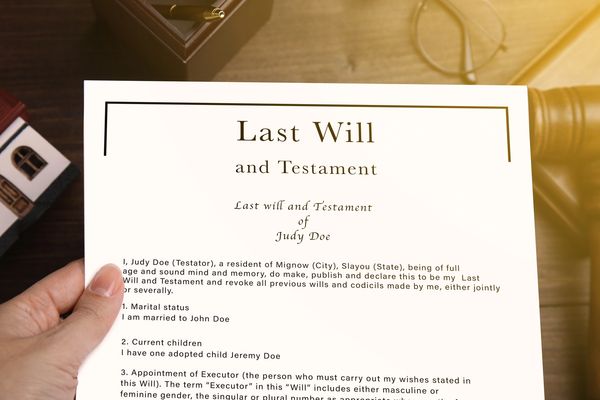 A "Last Will and Testament" document held by hands, with a miniature house in the background.
